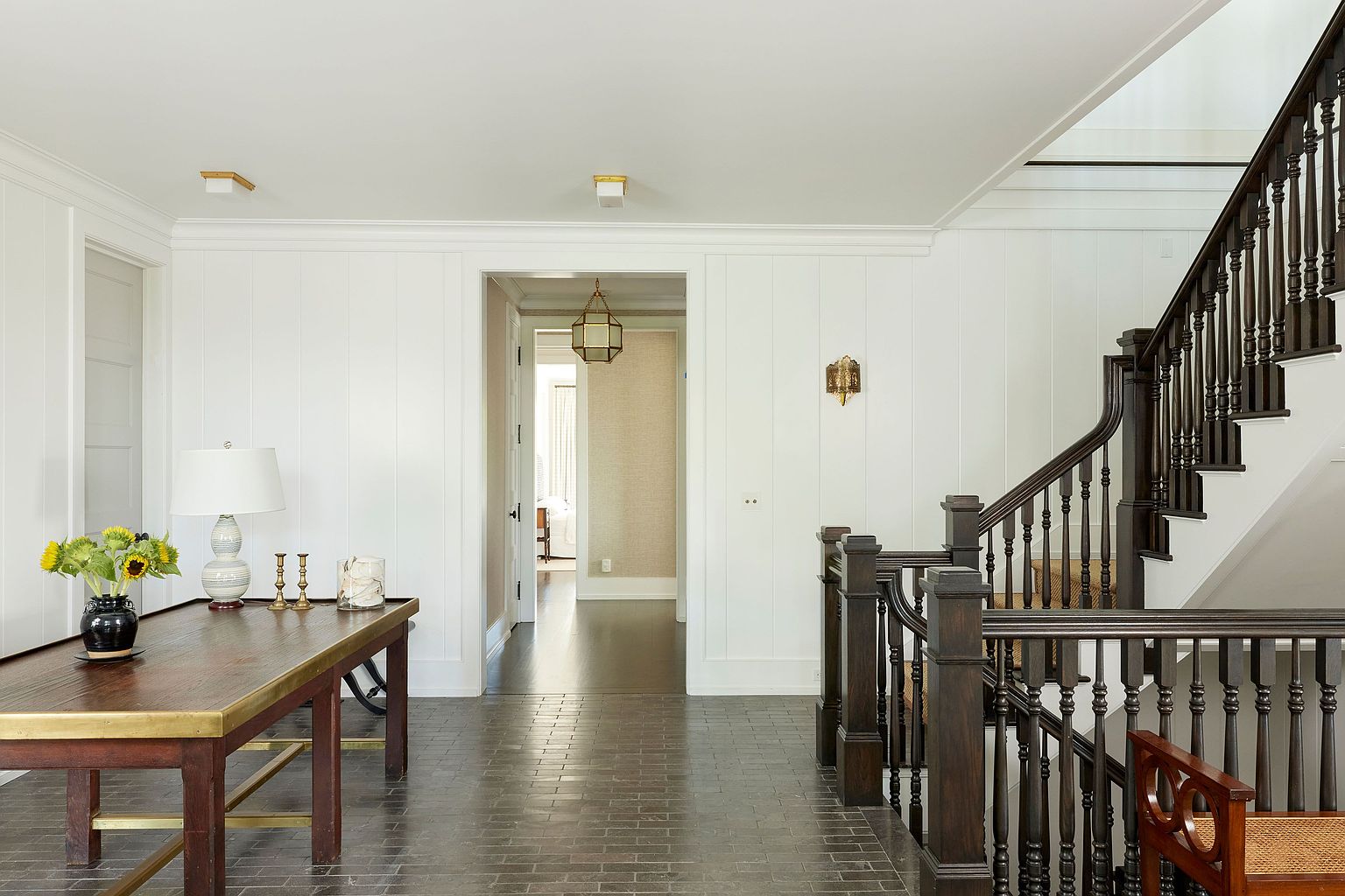 This interior shot showcases a well-lit hallway with dark wood stairs and railing, complemented by white paneled walls. A wooden table with brass accents sits to the left, adorned with a lamp and floral arrangement. The hallway leads to another room, creating a sense of depth and spaciousness, while the dark wood flooring adds a touch of elegance.
