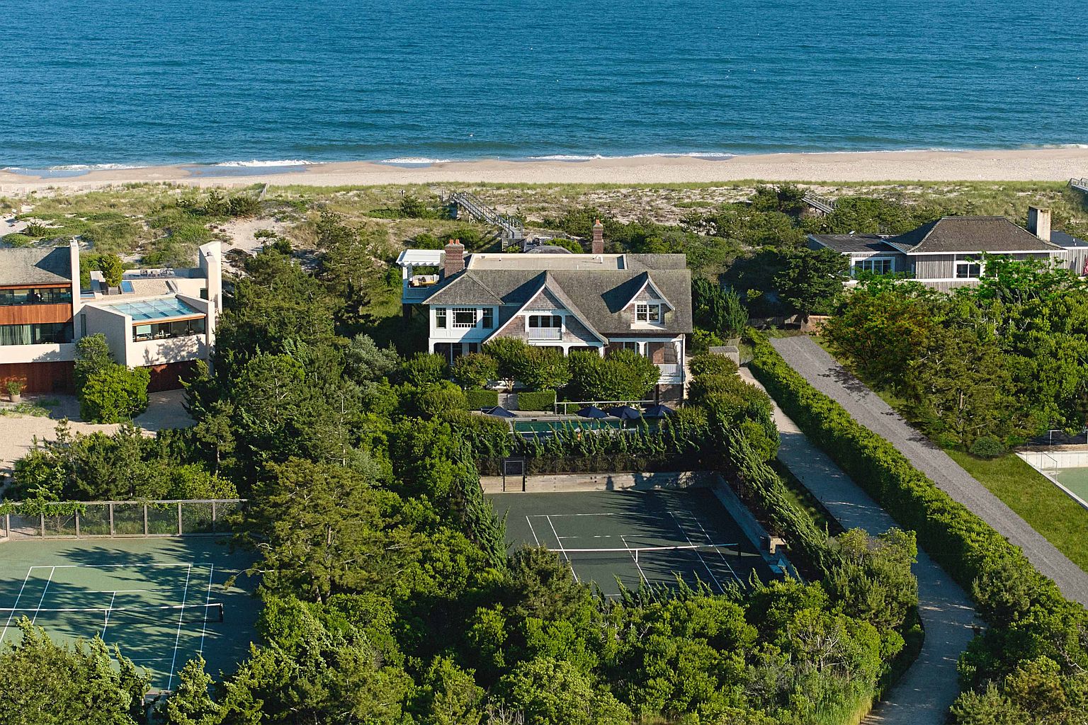 This aerial view showcases a luxurious beachfront property featuring a large house with a gray roof, a tennis court, and a swimming pool surrounded by lush greenery. The property is situated near a sandy beach and the ocean, offering stunning views and privacy. A long driveway leads to the house, and neighboring properties are also visible, all nestled within a landscape of trees and manicured hedges.