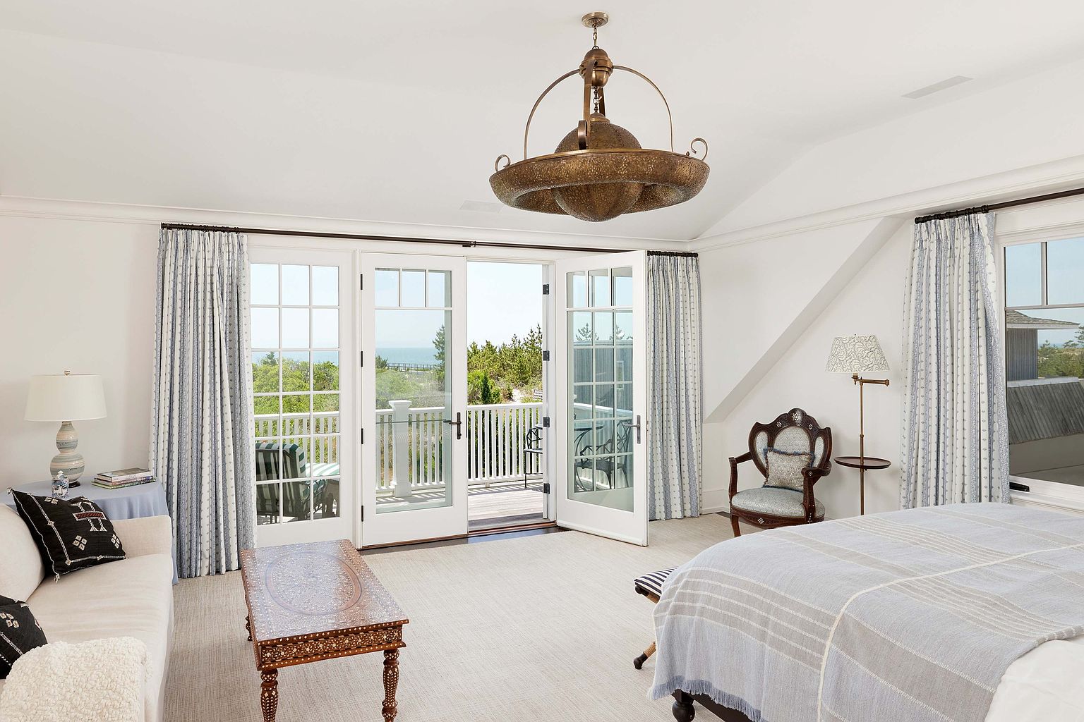 This primary bedroom features a light and airy atmosphere with white walls and large French doors leading to a balcony with ocean views. The room is furnished with a neutral-toned sofa, an ornate coffee table, and a bed with a blue and white striped throw. A unique chandelier hangs from the ceiling, adding a touch of elegance to the space.