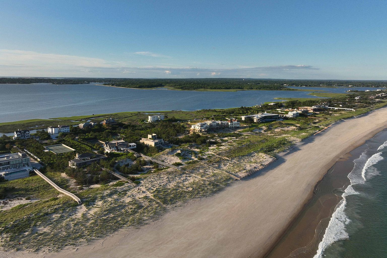 This aerial view showcases a stunning coastal property featuring luxurious homes nestled along a sandy beach. The scene includes a tranquil bay, lush greenery, and the expansive ocean, creating a picturesque and desirable setting. The perspective highlights the property's prime location and the serene lifestyle it offers.