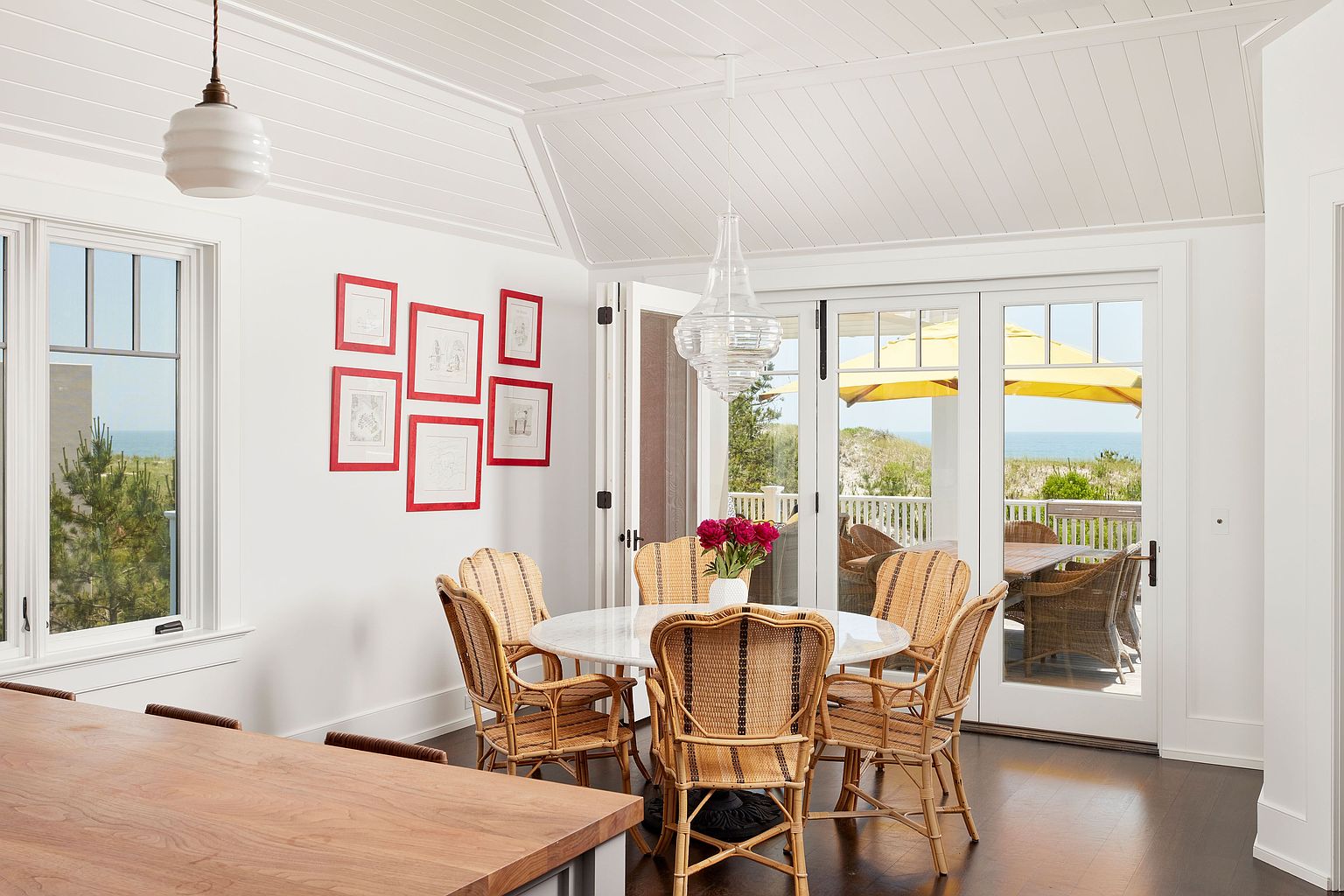 This is an interior shot of a dining room featuring a round marble-topped table surrounded by six wicker chairs. The room has white walls, a white planked ceiling, and dark hardwood floors. A set of framed artwork adorns one wall, and sliding glass doors lead to an outdoor patio area, offering a glimpse of the ocean.