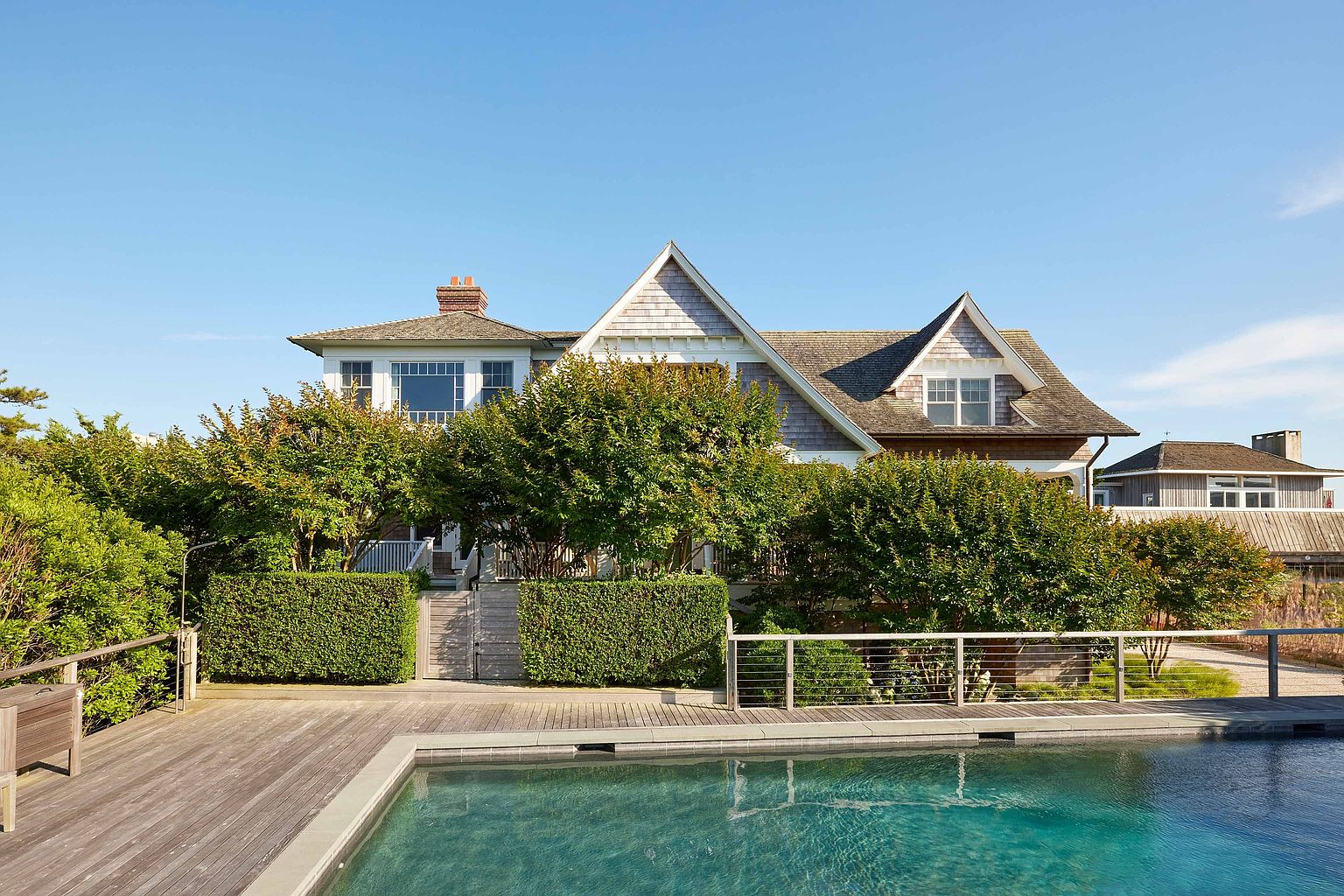 This image showcases a luxurious outdoor pool area with a wooden deck and a view of a large, well-maintained house. The pool's clear water invites relaxation, while the surrounding greenery and architectural details of the home create an upscale and private atmosphere. The scene suggests a high-end property perfect for entertaining and enjoying outdoor living.