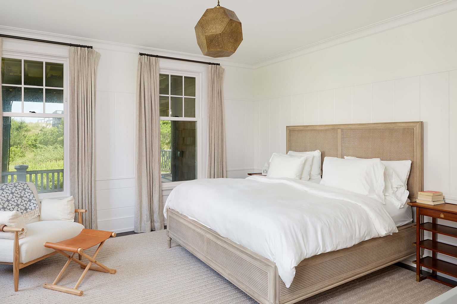 This is a bright and airy primary bedroom featuring a large bed with a woven headboard and white linens. Natural light floods the room through two large windows, complemented by neutral-toned curtains. A modern geometric pendant light hangs from the ceiling, adding a touch of elegance to the space.
