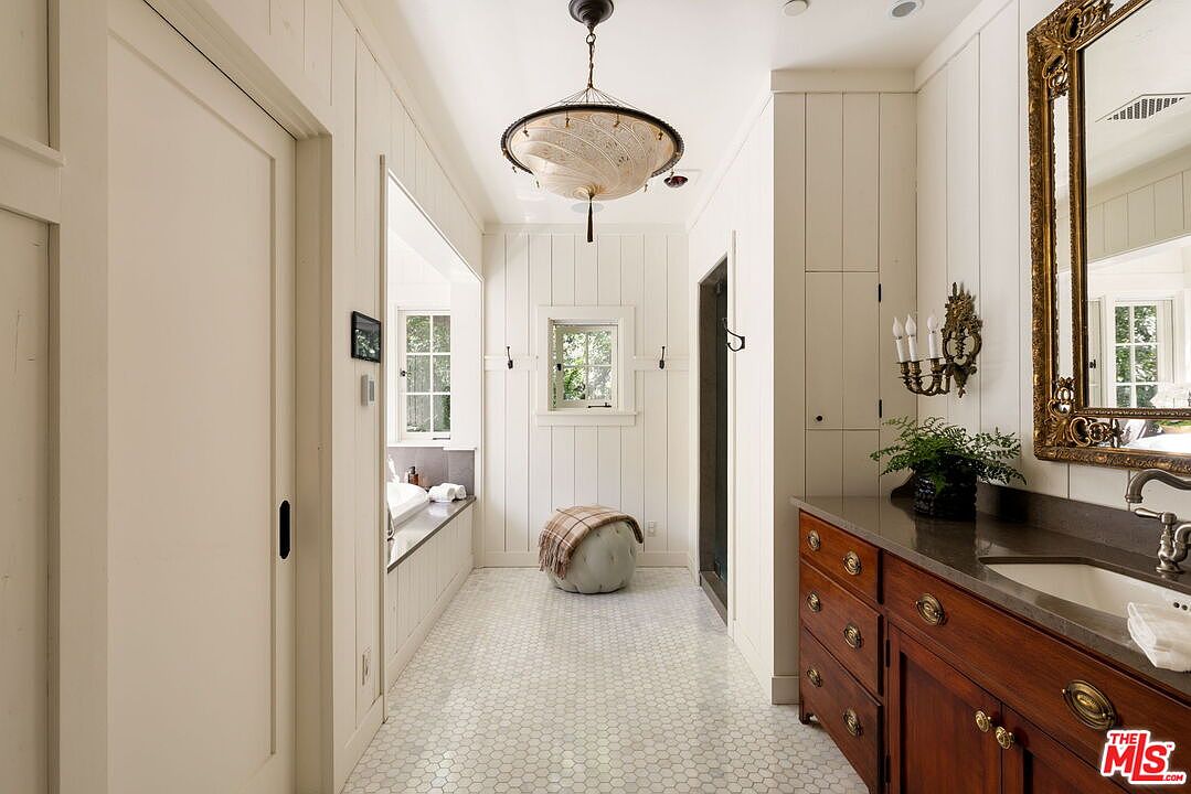 This elegant primary bathroom features a long hallway layout with white vertical paneling and classic penny-round tile flooring. A rich wood vanity with a dark stone countertop sits on the right, complemented by a decorative gold-framed mirror and wall-mounted sconces. At the end of the hall, a window provides natural light, while a unique, textured glass pendant light hangs centrally to create a sophisticated, spa-like atmosphere.