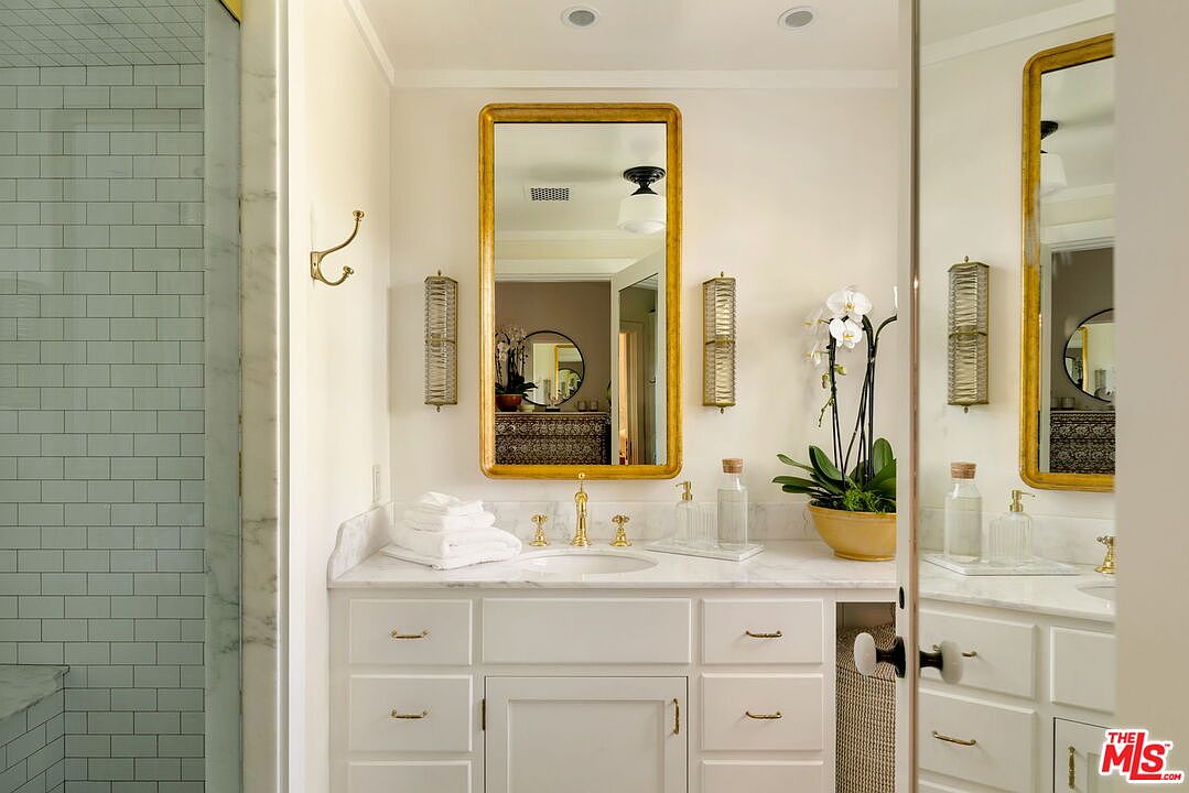 This bright and elegant bathroom features a white vanity topped with a light-colored stone counter and a gold-toned faucet. A large, gold-framed mirror is centered above the sink, flanked by two vertical wall sconces, while a potted orchid adds a touch of natural color to the space. To the left, a tiled shower area is visible, and the overall aesthetic is clean, sophisticated, and well-lit.