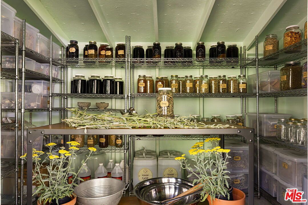 This organized pantry space features extensive wire shelving units filled with numerous glass jars containing dried herbs and ingredients. A central work table holds fresh herbs and metal mixing bowls, while potted yellow flowers add a touch of natural color to the foreground. The room is painted a soft green, creating a functional and rustic atmosphere for food preparation and storage.