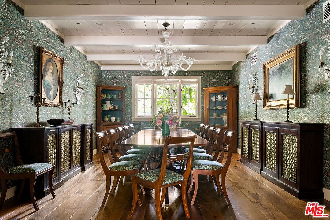 This formal dining room features a long wooden table surrounded by matching chairs, set against walls adorned with intricate floral wallpaper. A large crystal chandelier hangs from the white beamed ceiling, while two wooden cabinets flank a central window that brings in natural light. The room is anchored by dark wood sideboards with decorative metal mesh inserts, creating a sophisticated and traditional atmosphere.