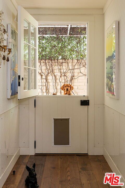 This charming hallway features a Dutch door that opens to reveal a view of a garden wall with climbing vines and a friendly dog looking in. The space is finished with light-toned walls, wood flooring, and framed artwork on either side, creating a warm and inviting atmosphere. A small decorative dog figurine sits on the floor in the foreground, adding a whimsical touch to this well-lit entry area.