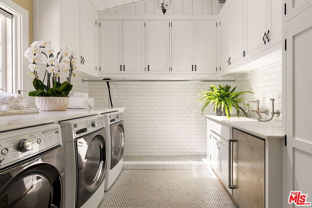 This bright and clean laundry room features white cabinetry, a subway tile backsplash, and a patterned tile floor. The space is equipped with a side-by-side washer and dryer set, a utility sink, and a hanging rod, all illuminated by under-cabinet lighting. The perspective is a straight-on shot looking down the length of the room, highlighting the organized and functional layout.