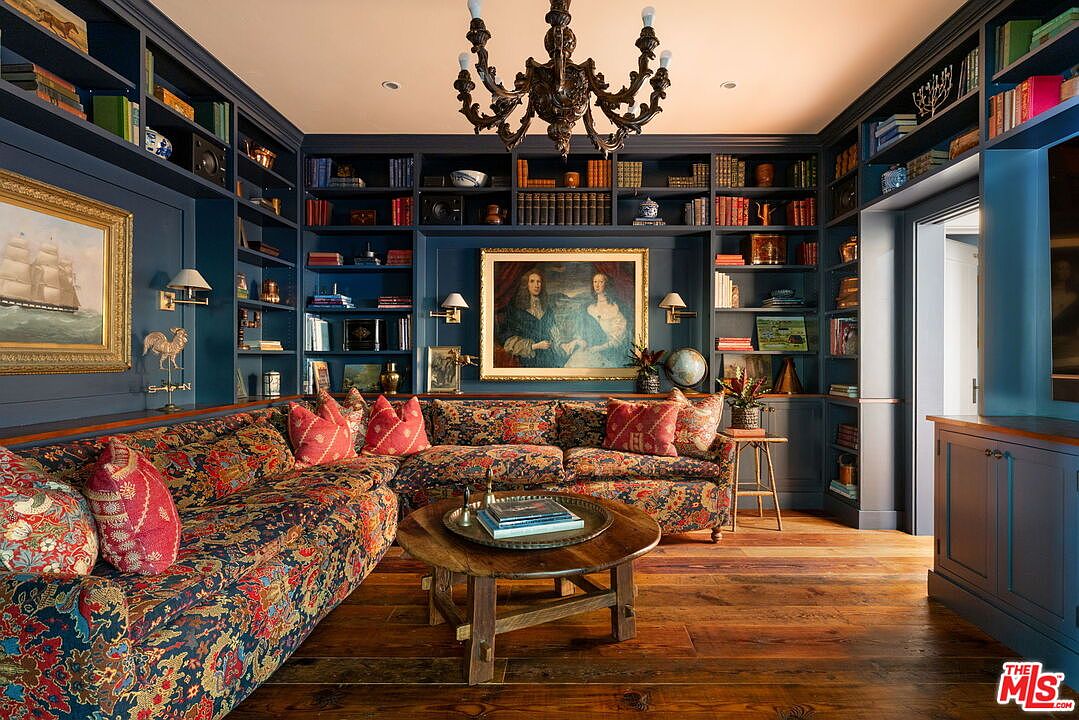 This sophisticated study features deep blue built-in shelving that spans the walls, creating a cozy and scholarly atmosphere. A large, patterned sectional sofa provides ample seating around a rustic wooden coffee table, while a classic chandelier hangs from the ceiling to provide warm lighting. The room is accented with traditional artwork and decorative objects, all set against rich wood flooring.
