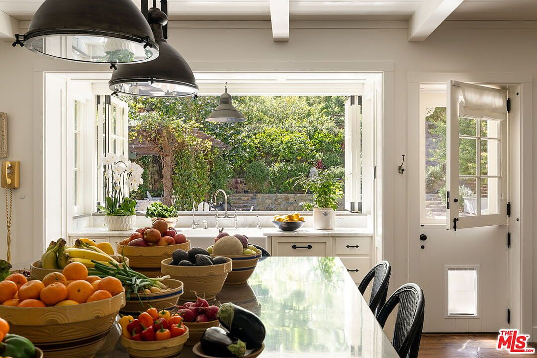 This bright and airy kitchen features a large central island topped with a reflective surface, abundantly stocked with fresh produce in rustic bowls. A wide, open window above the sink provides a seamless connection to the lush, green garden outside, while a Dutch door to the right adds a charming, traditional touch. The space is illuminated by large, industrial-style pendant lights and benefits from ample natural light, creating a warm and inviting atmosphere.