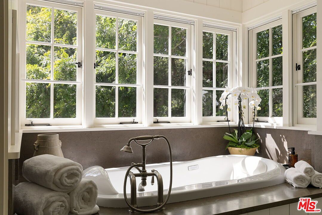 This serene primary bathroom features a deep soaking tub positioned beneath a series of large, multi-pane windows that flood the space with natural light. The room is accented by a bronze-toned faucet fixture, a stack of plush towels, and a potted orchid, creating a spa-like atmosphere. The neutral color palette and clean lines offer a sophisticated and tranquil aesthetic.
