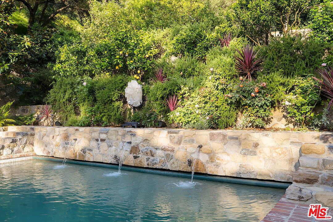This image captures a serene swimming pool area featuring a natural stone retaining wall with three integrated water spouts cascading into the water. The lush, terraced hillside behind the pool is densely planted with mature greenery, including lemon trees and ornamental grasses, and features a prominent, large white crystal sculpture as a focal point. The scene conveys a tranquil, resort-like atmosphere, emphasizing the integration of natural landscaping with custom water features.
