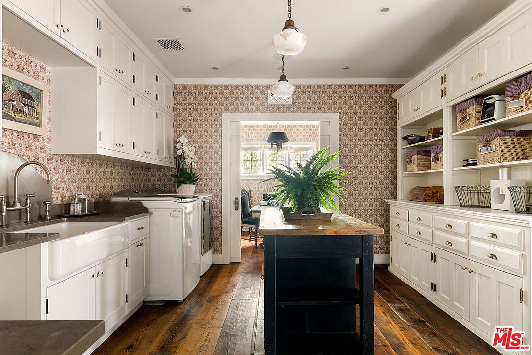 This charming laundry room features a classic farmhouse aesthetic with patterned wallpaper, white cabinetry, and a deep apron-front sink. A dark-toned central island provides additional workspace, while open shelving on the right offers convenient storage for baskets and household items. The room is anchored by warm-toned wood flooring and illuminated by two pendant lights, creating a cozy and functional space.