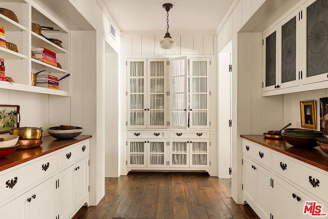 This elegant butler's pantry features custom white cabinetry with glass-fronted doors and dark hardware, providing ample storage and display space. The warm wood countertops contrast beautifully with the white woodwork, while the dark wood flooring adds a rich, grounded feel to the space. A classic pendant light hangs from the ceiling, illuminating the symmetrical layout that leads toward a central cabinet unit.