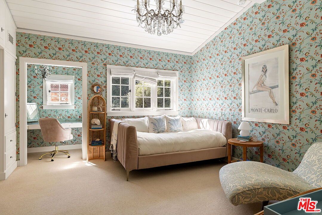 This charming guest bedroom features whimsical floral wallpaper, a plush daybed with decorative pillows, and a patterned chaise lounge. A crystal chandelier hangs from the white paneled ceiling, while a small alcove serves as a dedicated workspace with a desk and chair. The room is bright and airy, with large windows allowing natural light to fill the space, creating a cozy and inviting atmosphere.