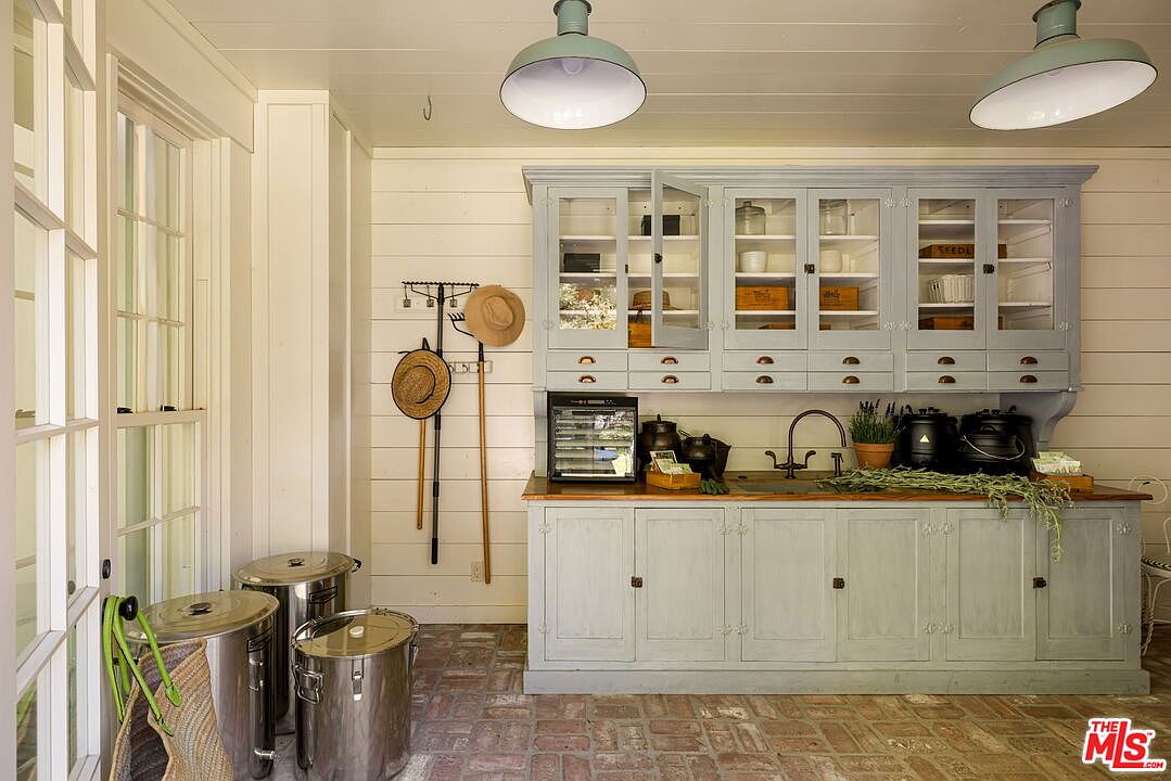 This charming, rustic-style potting room or kitchen workspace features light blue cabinetry with glass-front upper doors and a warm wood countertop. The space is accented by white shiplap walls, brick flooring, and two large, industrial-style pendant lights hanging from the ceiling. A sink is integrated into the counter, and the room includes practical storage for gardening tools, hats, and large metal canisters, creating a functional and aesthetic farmhouse atmosphere.