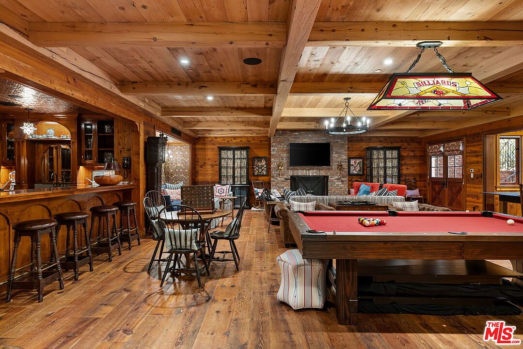 This expansive, rustic-style recreation room features warm wood-paneled walls and exposed ceiling beams, creating a cozy lodge-like atmosphere. The space is anchored by a large pool table under a stained-glass billiard light, with a stone-clad chimney breast housing a television above the fireplace in the background. A built-in bar area with stools and a small dining set complete the layout, making it an ideal space for entertainment and relaxation.