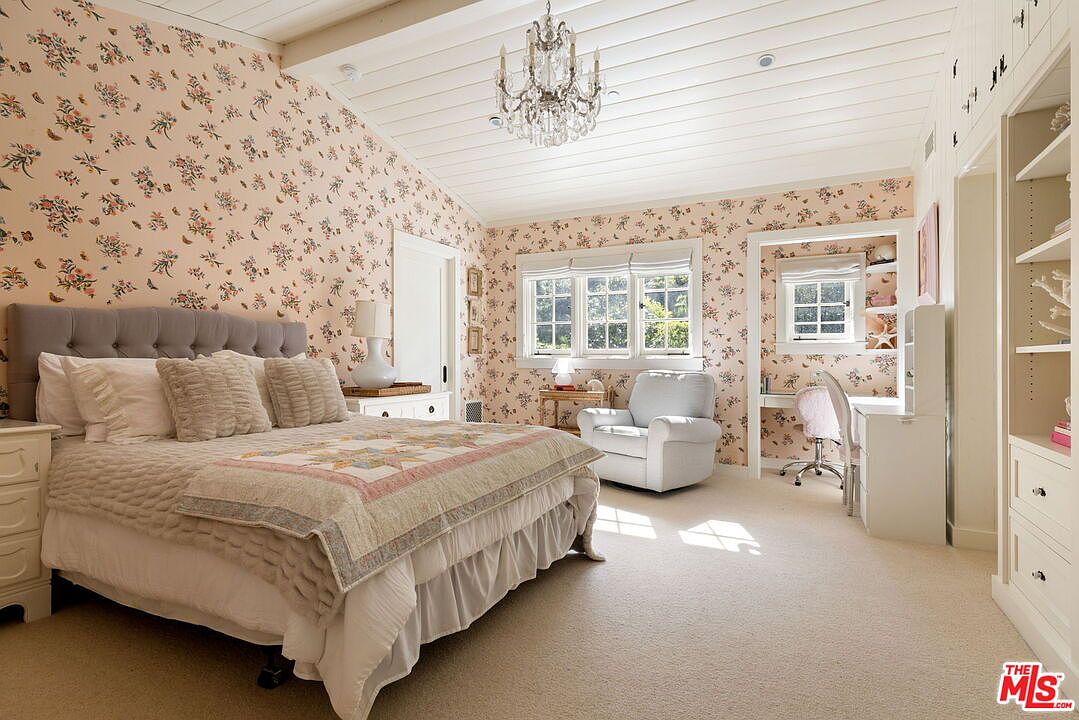 This charming bedroom features a vaulted ceiling with white shiplap paneling and a classic crystal chandelier. The walls are adorned with a delicate floral wallpaper, complemented by a tufted headboard and a cozy quilt on the bed. A built-in desk area and shelving unit provide functional workspace, while a comfortable armchair sits near the windows, creating a bright and inviting atmosphere.