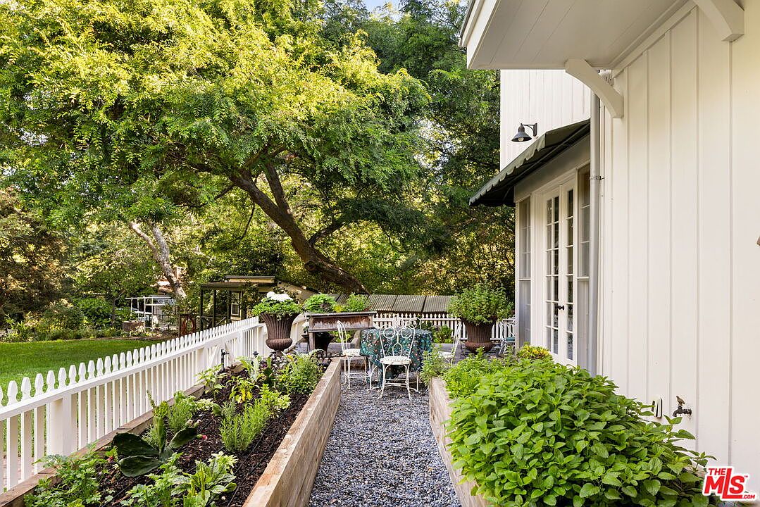 This charming outdoor space features a gravel pathway flanked by raised garden beds filled with lush greenery and herbs. A white picket fence separates the garden from a grassy lawn, while a small bistro set sits on the gravel, creating an inviting spot for relaxation. The scene is framed by a white-sided house with a dark awning and mature trees that provide a canopy of shade over the backyard.