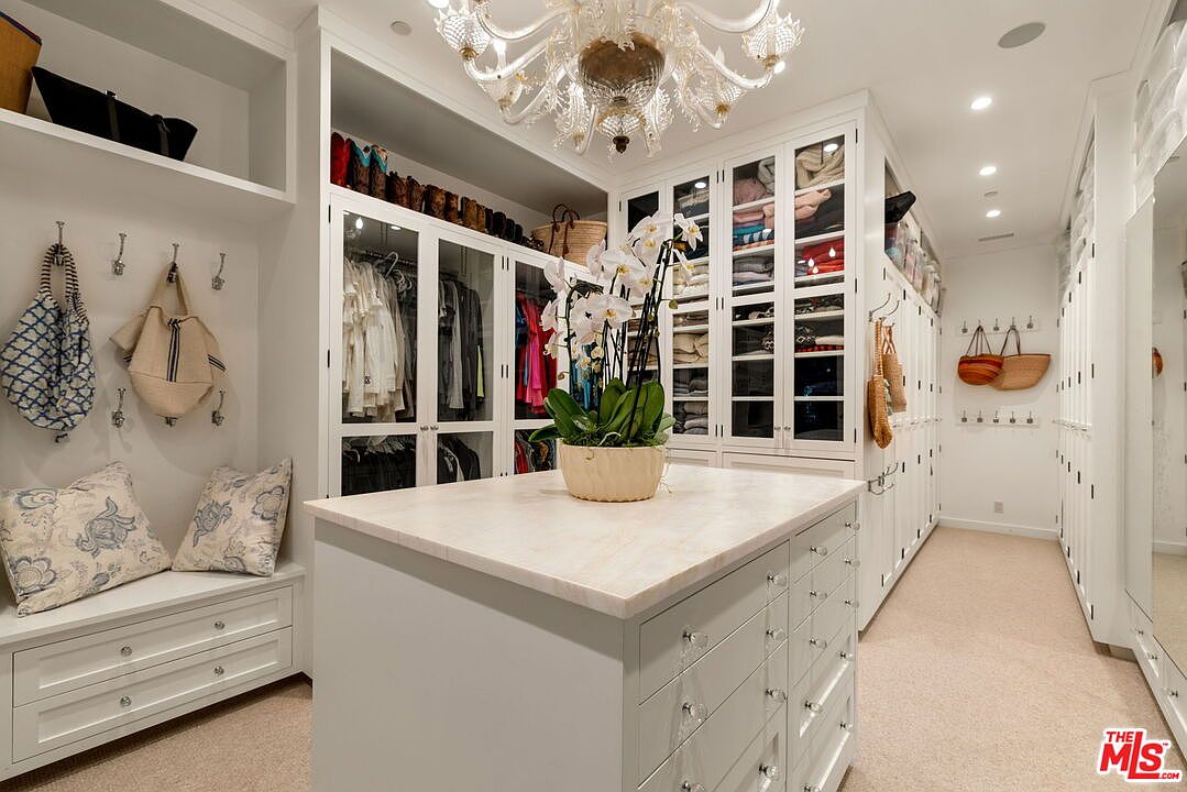 This spacious walk-in closet features a central island with a light-toned stone countertop and ample drawer storage. The room is lined with white cabinetry, including glass-fronted doors for display and open shelving for accessories, all illuminated by a decorative chandelier. A built-in bench with patterned pillows and wall-mounted hooks provides a functional seating and storage area, creating an organized and luxurious atmosphere.