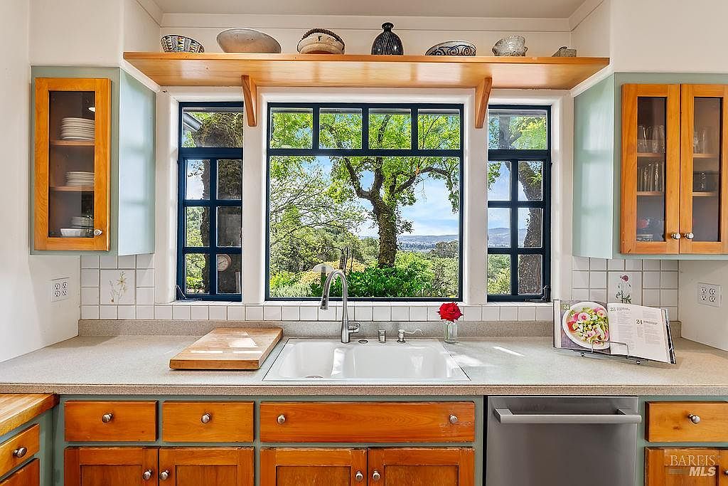 This kitchen features a bright, functional workspace centered around a large window that frames a scenic outdoor view. The space includes light-toned wood cabinetry with contrasting painted frames, a white double-basin sink, and a neutral countertop. A wooden shelf above the window provides display space for decorative items, while a cookbook rests on the counter, adding a lived-in, inviting feel to the room.