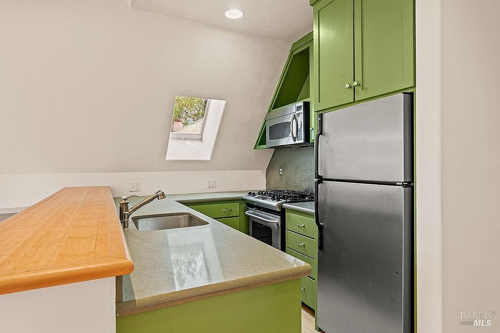 This kitchen features a unique sloped ceiling with a skylight that brings in natural light. The space is equipped with green cabinetry, a stainless steel refrigerator, and a built-in microwave, complemented by a light-toned countertop and a wood-topped breakfast bar. The perspective captures the compact, functional layout of the cooking area from the vantage point of the adjacent living space.