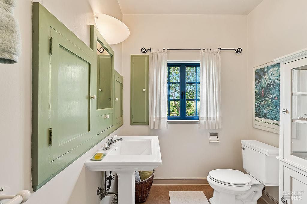 This charming bathroom features a classic pedestal sink paired with a unique, sage-green custom cabinetry unit that incorporates a mirror and storage. A window with blue frames and white curtains allows natural light to brighten the space, which also includes a white toilet and a tall storage cabinet. The room has a clean, vintage-inspired aesthetic with a neutral color palette.