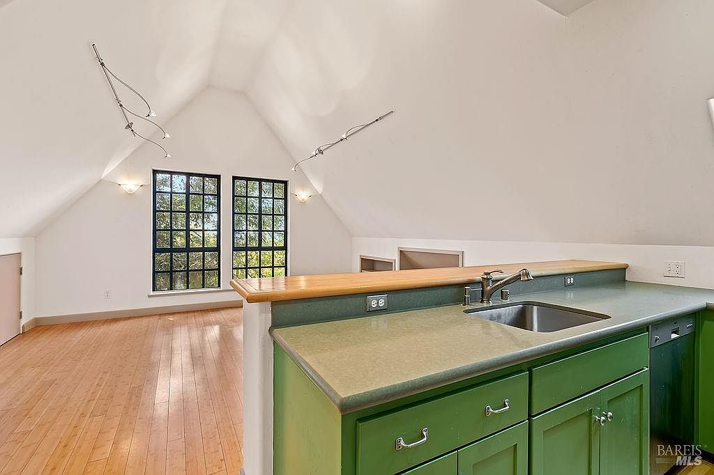 This interior view captures a kitchen area with green cabinetry and a light-toned countertop, featuring a stainless steel sink and faucet. The kitchen opens into a spacious, vaulted-ceiling room with light wood flooring and large windows that allow for ample natural light. The perspective is from the kitchen counter looking out into the adjacent living space, highlighting the open-concept layout of the home.