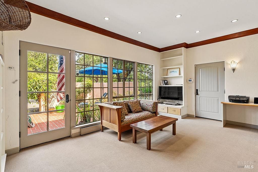 This bright living room features neutral carpeting and a comfortable seating area centered around a wooden coffee table. Large windows and a glass-paned door provide ample natural light and a view of the outdoor patio, while built-in shelving surrounds a television console. The space is designed for relaxation, with a clean, functional layout that seamlessly connects to the exterior.
