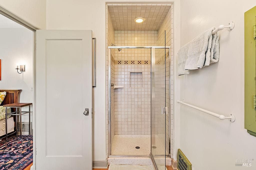 This bathroom features a walk-in shower enclosed by a glass door, finished with light-colored square tiles and a recessed niche. To the left, a white door leads into an adjacent bedroom, while a towel rack and a wall-mounted bar are positioned on the right wall. The space is illuminated by a recessed ceiling light, creating a clean and functional aesthetic.
