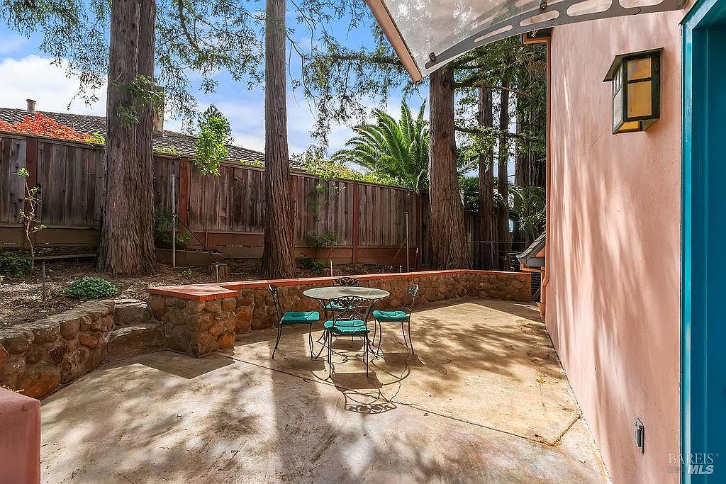This image captures a private, shaded patio area featuring a concrete surface and a small bistro set with four chairs. The space is defined by a low, rustic stone retaining wall that separates the patio from a wooded backyard filled with tall trees and a wooden fence. The perspective is from the side of a pink-toned house, looking out toward the garden, creating a quiet and secluded outdoor retreat.