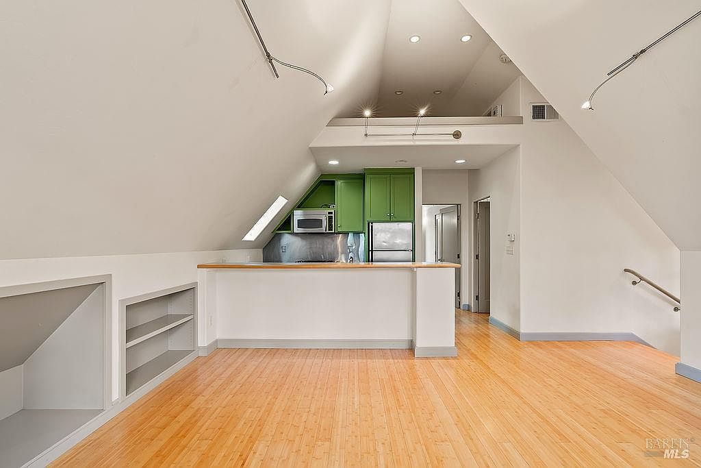 This interior view captures a kitchen area set within an attic-style space featuring dramatic sloped ceilings and light wood flooring. The kitchen is defined by a breakfast bar with a light wood countertop, vibrant green cabinetry, and stainless steel appliances, including a microwave and refrigerator. The perspective is from the living area looking toward the kitchen, highlighting the open layout and the unique architectural geometry of the room.