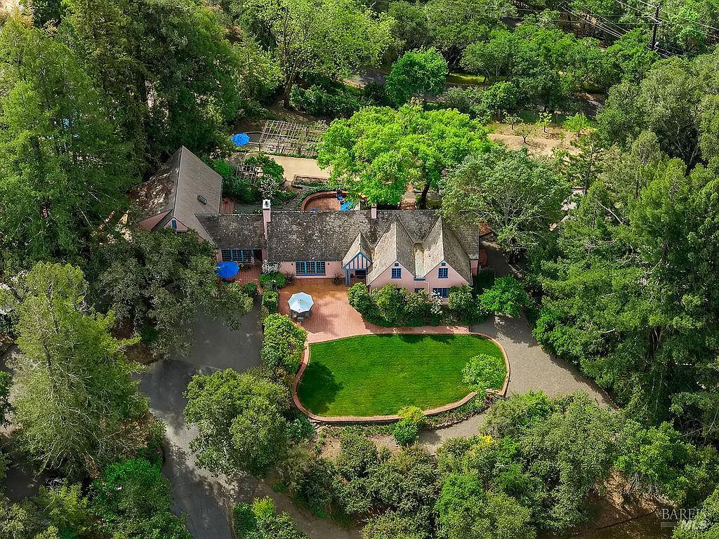 This high-angle aerial view captures a charming, multi-gabled residence nestled within a lush, tree-filled landscape. The property features a brick-paved patio area, a manicured oval lawn, and a private swimming pool tucked behind the main structure. The home's traditional architecture is complemented by mature greenery and winding pathways, creating a secluded and serene estate atmosphere.
