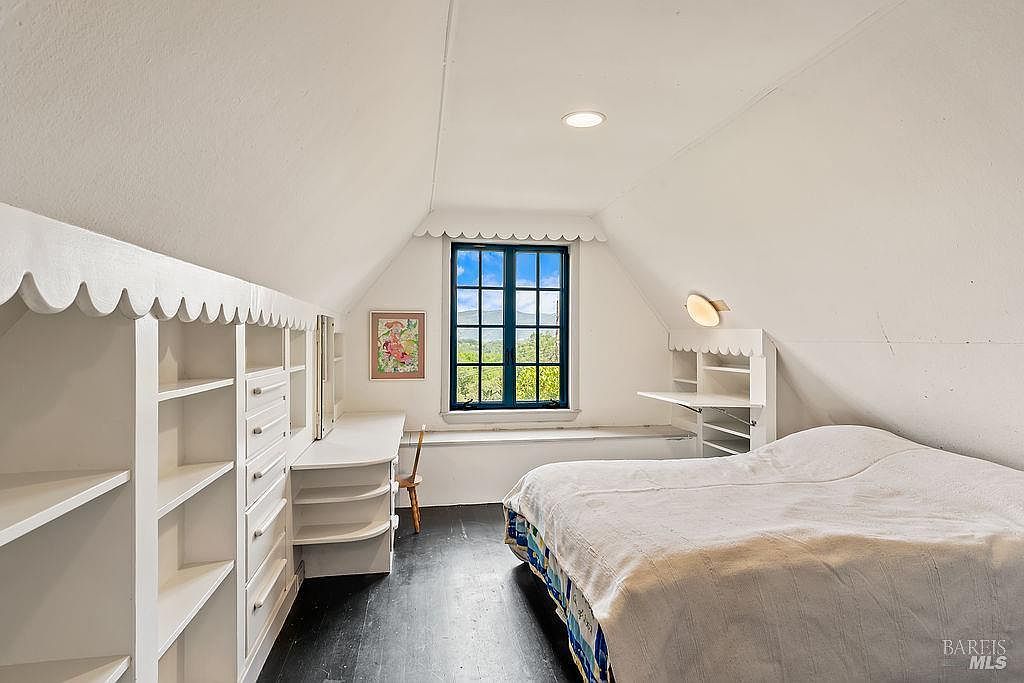 This cozy attic bedroom features a sloped ceiling, dark wood flooring, and a built-in white storage unit with integrated shelving and a desk area. A central window with dark blue trim provides a scenic view of the outdoors, while a bed with a light-colored quilt occupies the right side of the room. The space has a charming, cottage-like aesthetic with scalloped trim details along the built-in furniture.