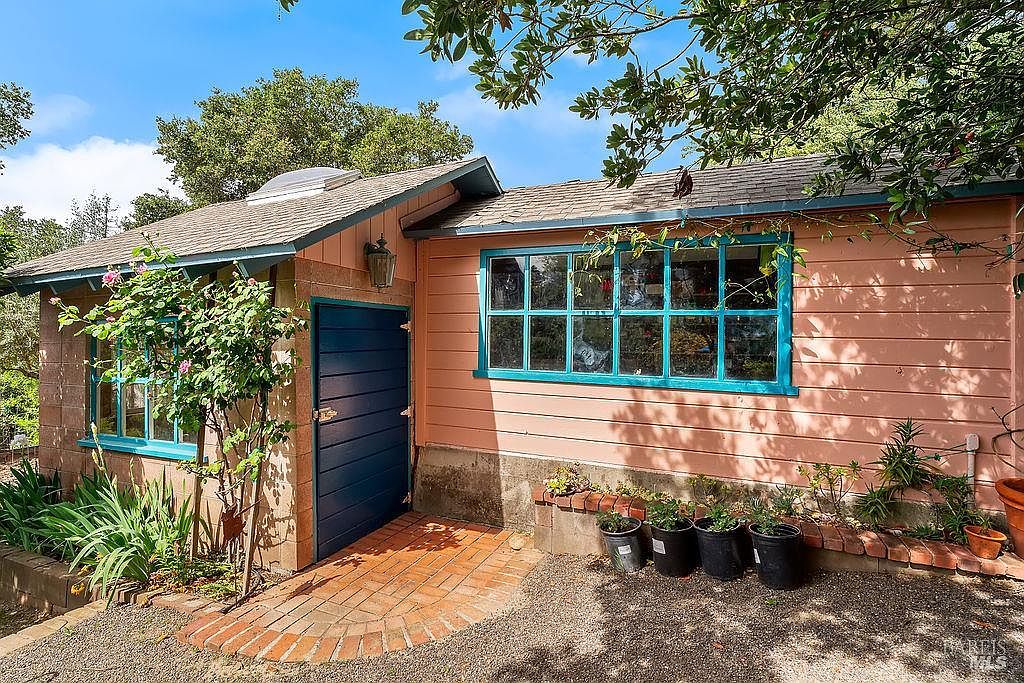 This side view of a charming, single-story home features light-toned horizontal siding accented by a vibrant blue door and matching window frames. A small brick-paved entryway leads to the door, complemented by a low brick planter box filled with potted plants. The scene is framed by lush greenery and tree branches, creating a peaceful and inviting atmosphere.