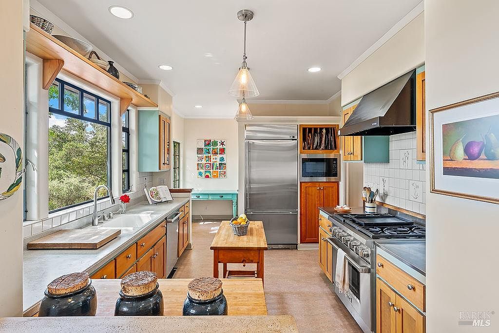 This bright and inviting kitchen features a blend of light wood cabinetry, a central wooden island, and a large window above the sink that provides ample natural light. The space is equipped with stainless steel appliances, including a professional-style range with a dark hood and a large refrigerator, complemented by white tiled backsplashes. A small, colorful art piece hangs on the wall near a light blue console table, adding a touch of character to the functional and well-organized layout.