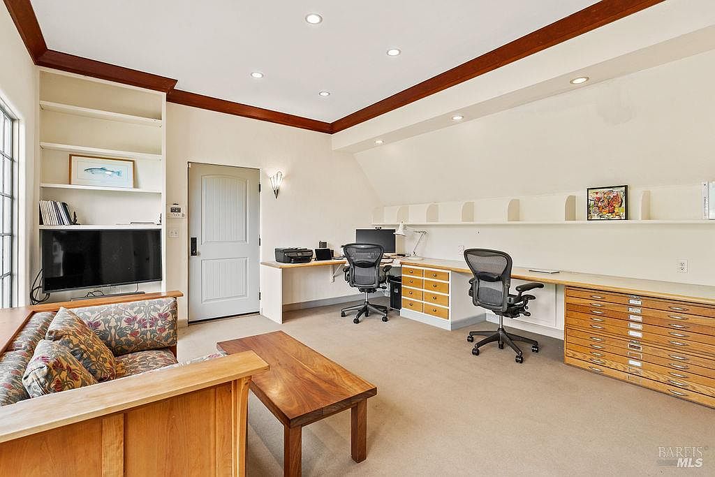 This spacious home office features a dual-desk workstation with built-in storage drawers and extensive shelving along the wall. A comfortable seating area with a patterned sofa and a wood coffee table is positioned in the foreground, creating a versatile multi-use space. The room is illuminated by recessed lighting and features neutral carpeting, providing a professional yet inviting atmosphere.