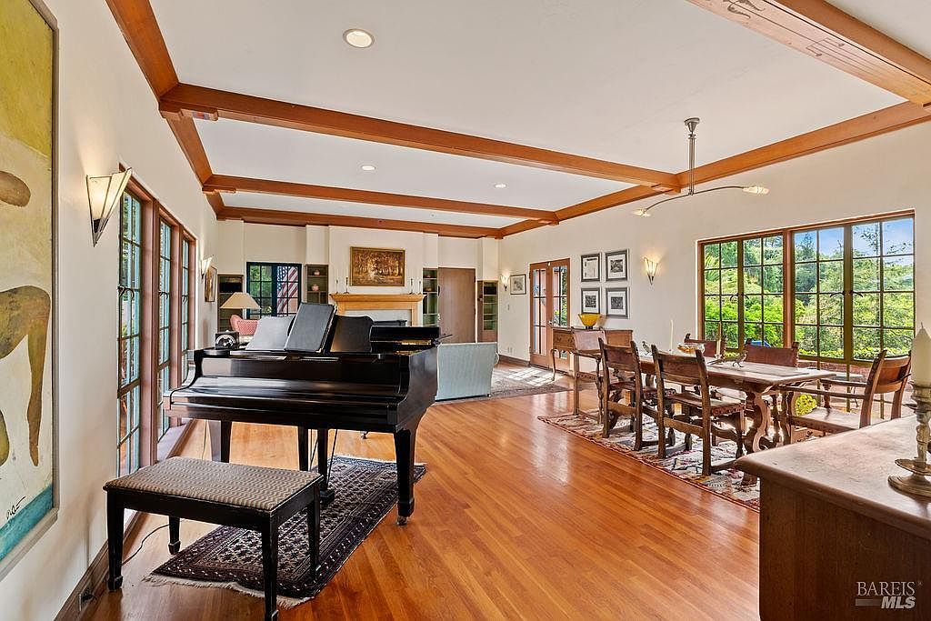 This spacious, open-concept living and dining area features warm wood flooring and exposed wooden ceiling beams that add architectural character. A grand piano serves as a focal point in the foreground, while a dining set sits near large windows that provide abundant natural light and views of the surrounding greenery. The room is decorated with framed artwork and wall sconces, creating an elegant and inviting atmosphere.