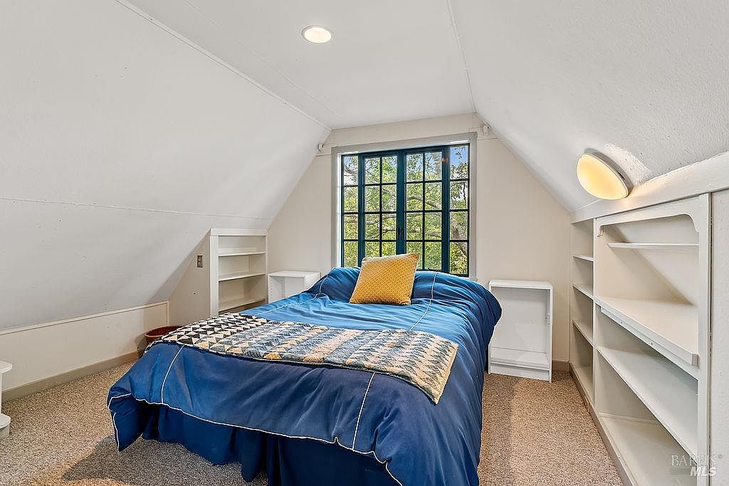 This cozy attic bedroom features sloped ceilings and a central window that provides natural light. The room is furnished with a bed dressed in a blue comforter and a yellow accent pillow, flanked by built-in shelving units on either side. The space has a neutral color palette and carpeted flooring, creating a comfortable and functional living area.