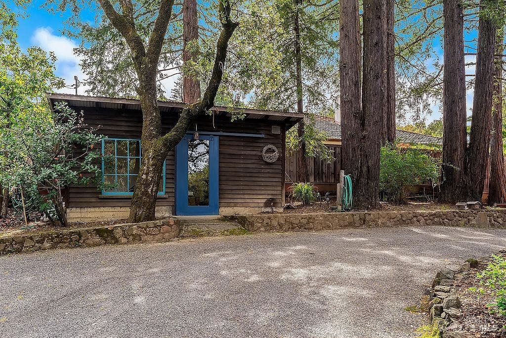 This rustic, wood-sided structure features a distinctive blue door and matching window frames, set against a backdrop of towering redwood trees. A gravel driveway leads up to the building, which is bordered by a low stone retaining wall. The scene captures a serene, woodland setting with a natural, cabin-like aesthetic.