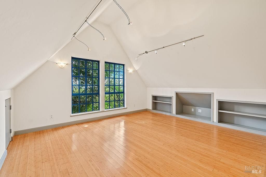 This bright attic space features vaulted ceilings and light wood flooring, creating an airy and open atmosphere. Two large windows with grid detailing allow for ample natural light, while built-in shelving units provide functional storage along the sloped walls. The room is illuminated by track lighting mounted on the ceiling, offering a versatile area suitable for a bedroom, office, or hobby space.