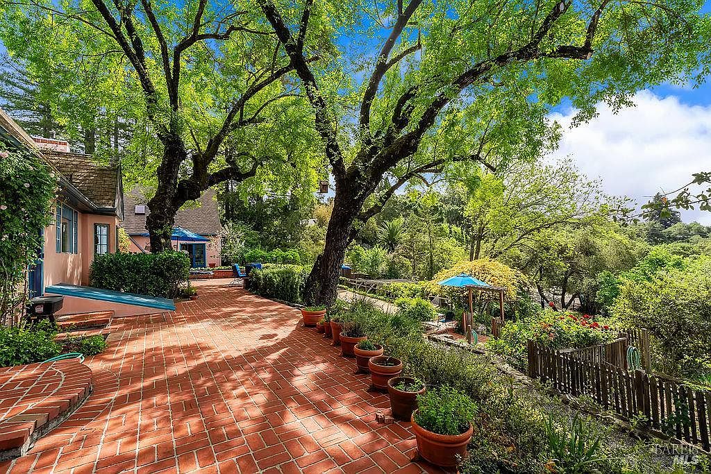 This inviting outdoor patio features classic red brick pavers and is shaded by a large, mature tree, creating a serene and private atmosphere. The space is lined with numerous potted plants and overlooks a lush, green garden area with a wooden fence and a small blue umbrella. The perspective captures the transition from the home's exterior wall to the expansive, sun-dappled terrace, highlighting a peaceful retreat perfect for relaxation.
