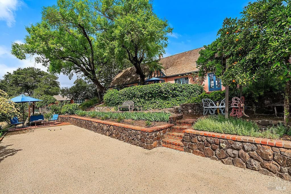 This expansive outdoor space features a gravel bocce ball court bordered by a rustic stone retaining wall with brick capping. A lush, manicured hedge separates the lower court from the elevated house, which is framed by mature trees and a vibrant orange tree. The scene is captured from a low angle, emphasizing the tiered landscaping and the inviting, sun-drenched atmosphere of the property.