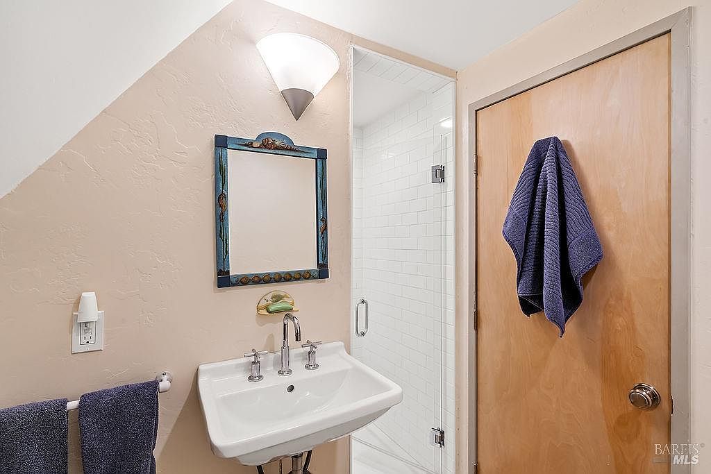 This compact bathroom features a wall-mounted white pedestal sink with a chrome faucet and a decorative framed mirror above. To the right, a glass-enclosed shower stall is lined with white subway tiles, while a wooden door with a blue towel hanging on it provides access. The room is illuminated by a modern wall-mounted light fixture, creating a clean and functional space.