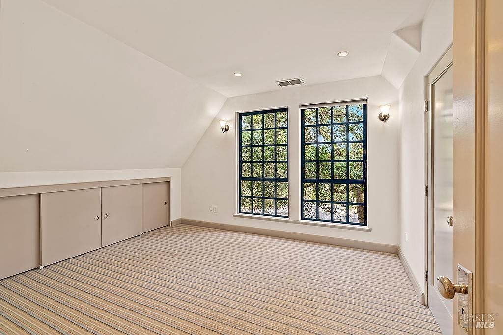 This bright, carpeted room features a sloped ceiling and built-in storage cabinets along the lower wall. Two large, multi-pane windows allow ample natural light to fill the space, offering a view of the trees outside. The room is finished with neutral walls and wall-mounted light fixtures, creating a clean and versatile living area.