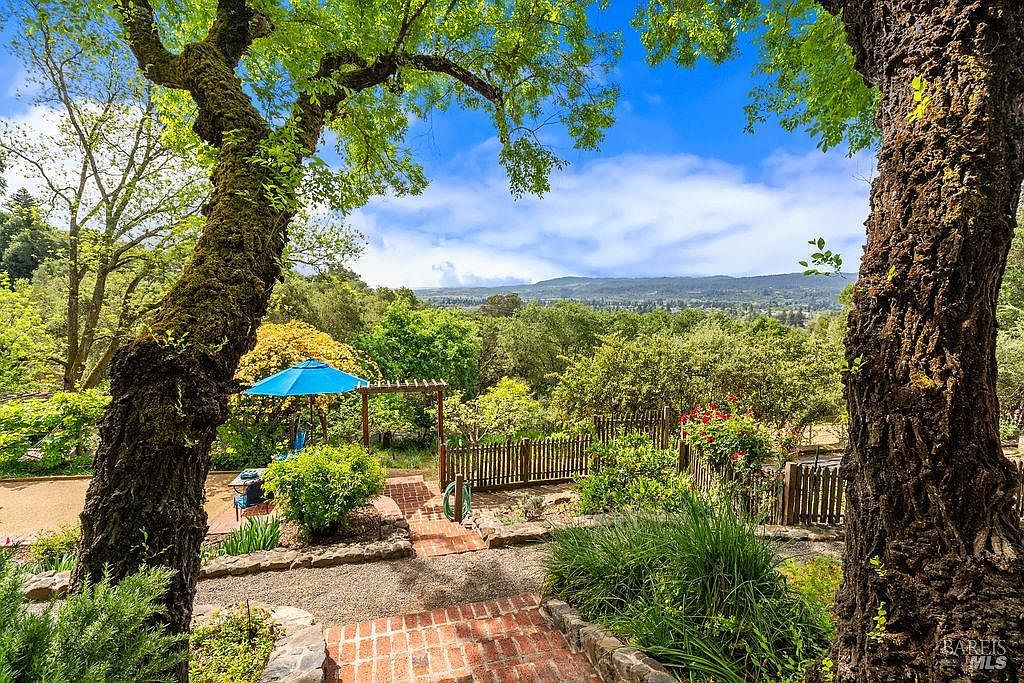 This scenic outdoor space features a brick-paved path leading through a lush, terraced garden framed by two large, textured tree trunks in the foreground. A blue umbrella provides shade over a small seating area, while a wooden pergola and a rustic picket fence define the garden's boundaries. The background offers a sweeping, elevated view of rolling green hills and a distant valley under a bright, clear sky.
