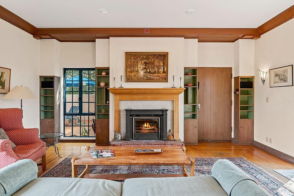 This cozy living room features a central fireplace with a wood mantel and a framed landscape painting above it, flanked by built-in shelving units. The room is furnished with a patterned armchair and a light-colored sofa, centered around a wooden coffee table on a large, ornate area rug. A large window to the left of the fireplace provides a view of an outdoor patio, while warm wood trim accents the ceiling and flooring throughout the space.
