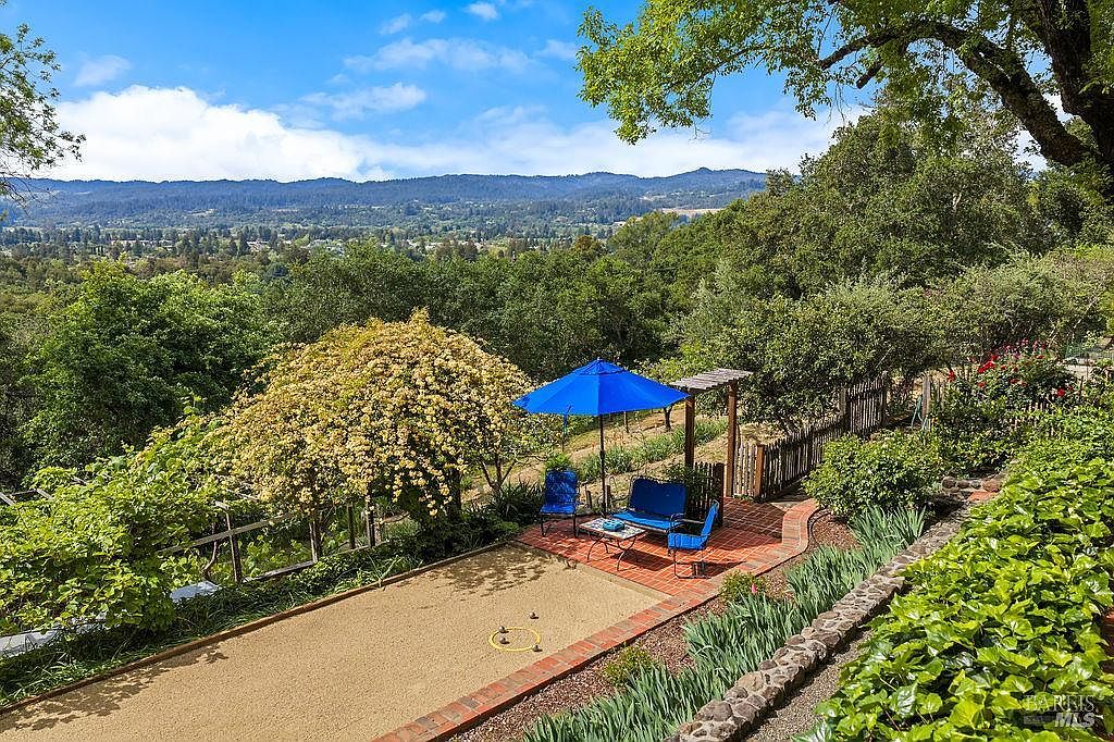 This elevated outdoor space features a bocce ball court set against a backdrop of rolling hills and lush greenery. A brick-paved patio area includes a blue umbrella, outdoor seating, and a wooden arbor, creating a serene spot for relaxation. The scene is framed by mature trees and vibrant landscaping, offering a picturesque view of the surrounding valley.
