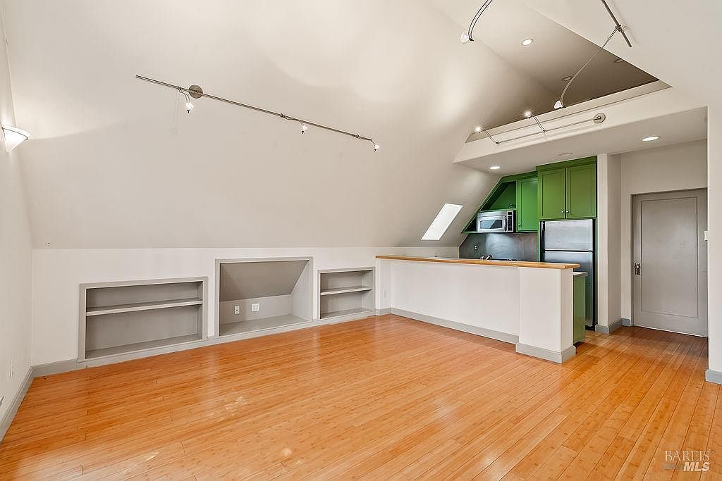 This open-concept living area features light wood flooring and a dramatic vaulted ceiling that follows the roofline. The space includes built-in shelving units along the lower wall and a breakfast bar that separates the living area from a compact kitchen with green cabinetry. A skylight provides natural illumination, while track lighting mounted on the angled ceiling offers additional brightness to the room.